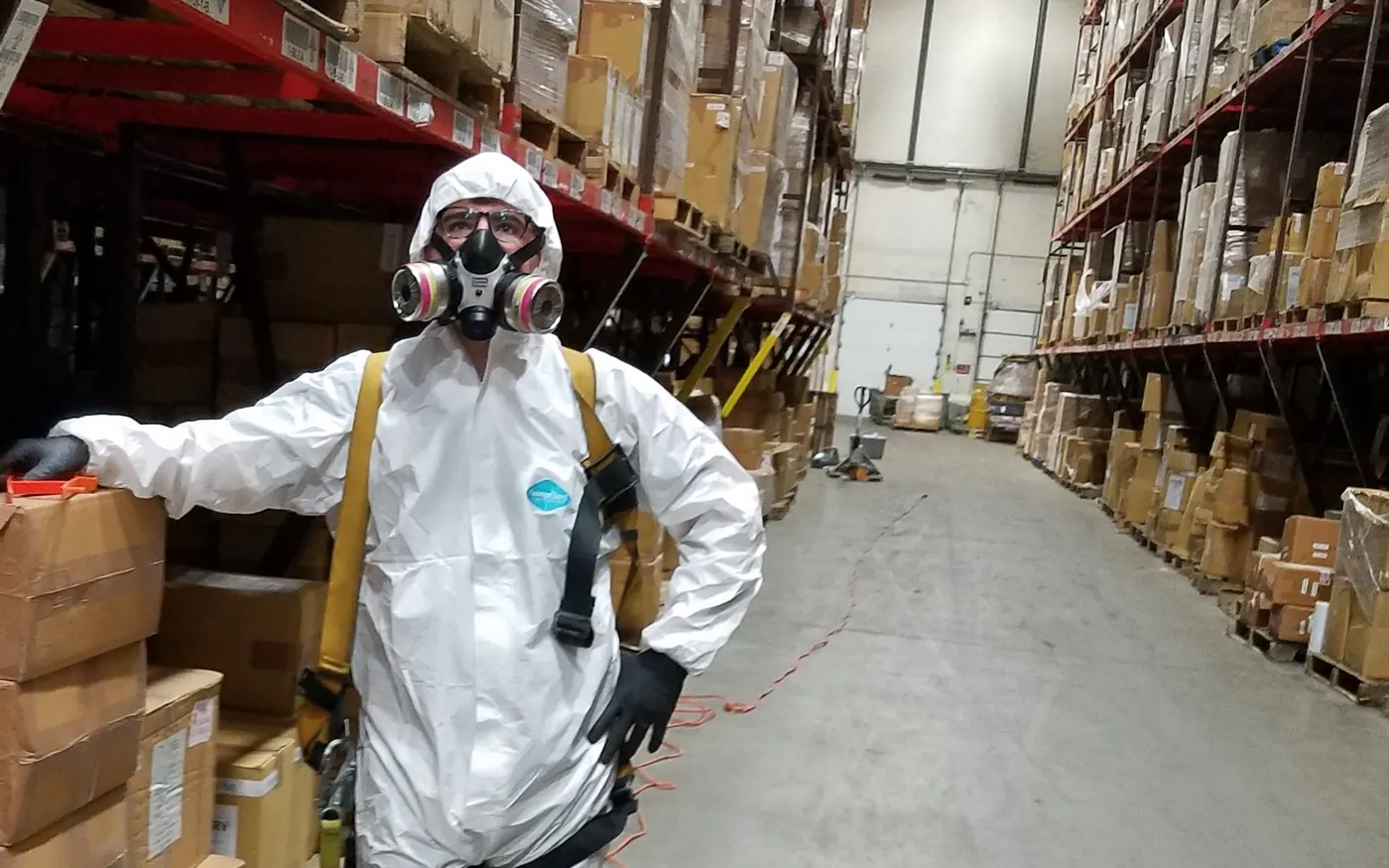 A Marrone technician in a Tyvek suit and respirator standing in a warehouse aisle between racks of stacked pallets