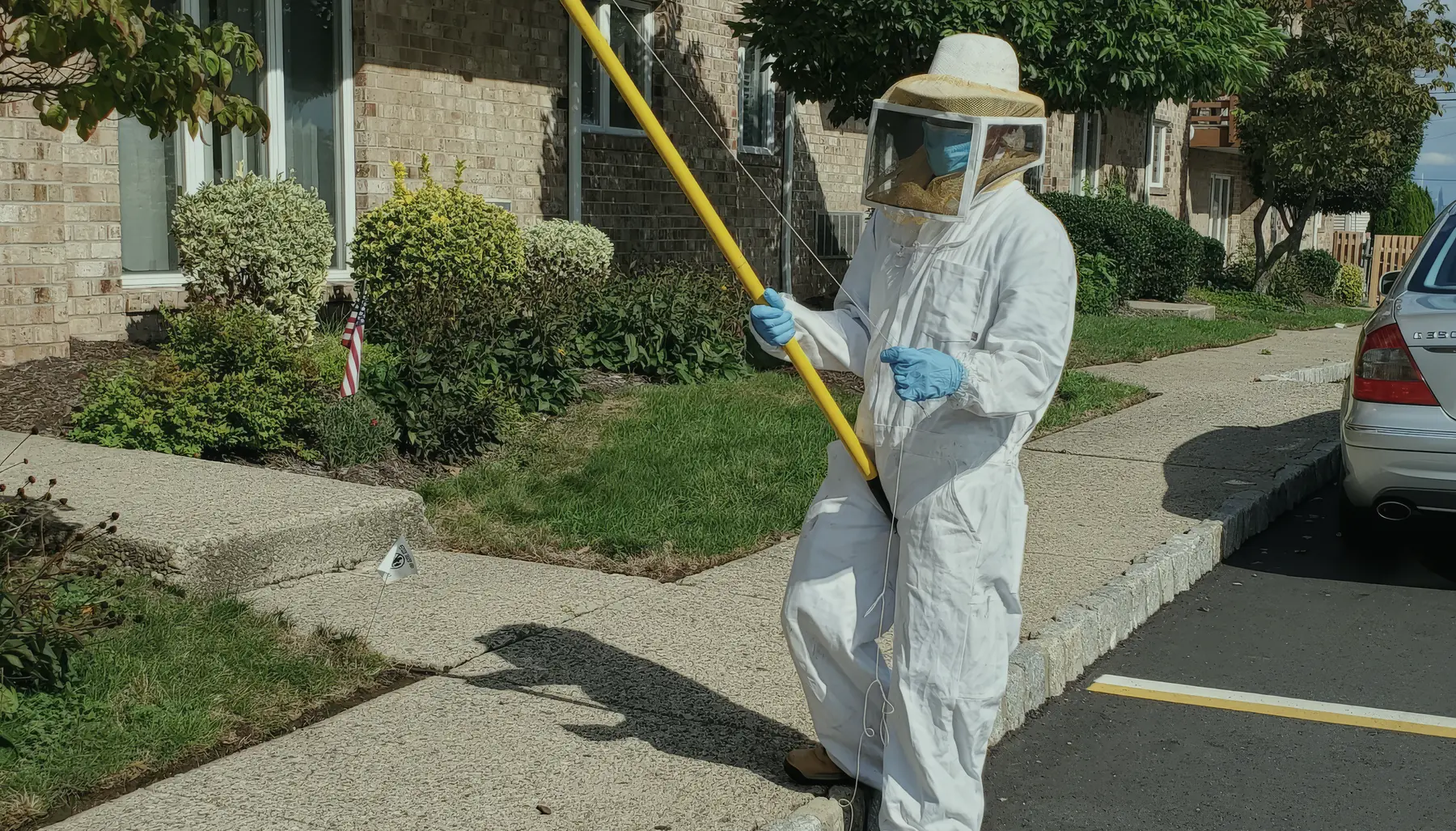 A Marrone Pest Management technician in a white bee suit and veil, holding a long yellow pole sprayer up to the balcony soffit of a brick apartment building on a bright day