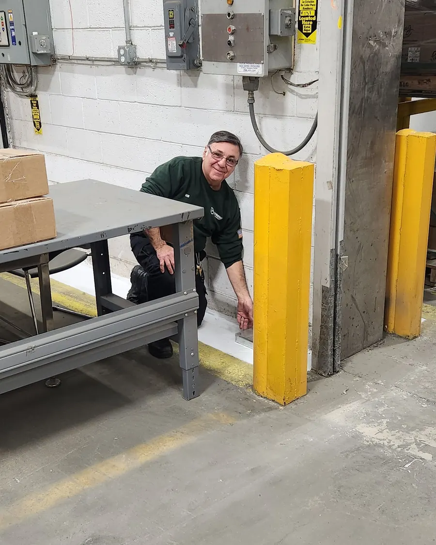Ron Marrone, President, kneeling at a bait station beside a loading dock in a food distribution warehouse