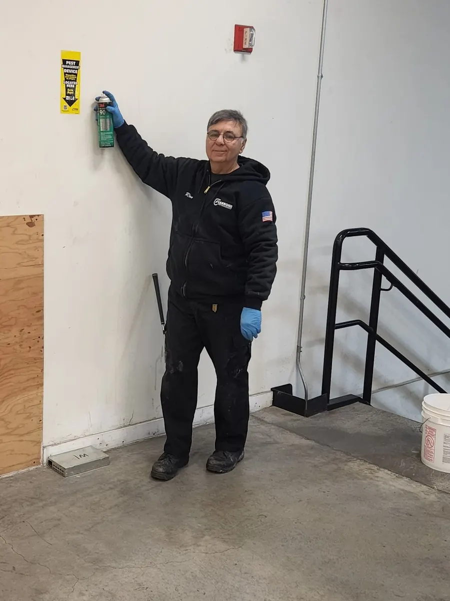 A Marrone technician treating a yellow pest monitoring device mounted on a warehouse wall with an aerosol applicator