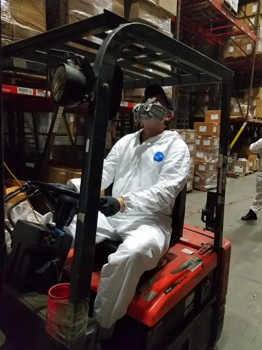A Marrone technician in a white Tyvek suit and respirator seated on a forklift in a warehouse aisle, looking up at the racking